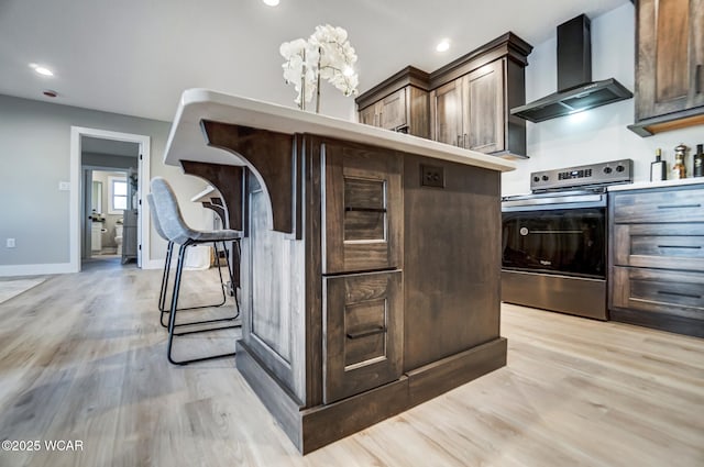 kitchen with dark brown cabinetry, wall chimney exhaust hood, light wood-type flooring, a kitchen breakfast bar, and stainless steel electric stove