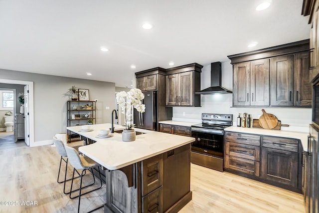 kitchen featuring wall chimney exhaust hood, light hardwood / wood-style flooring, appliances with stainless steel finishes, a kitchen breakfast bar, and a kitchen island with sink