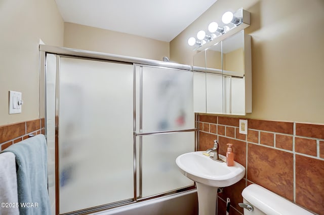 full bath featuring tile walls, toilet, combined bath / shower with glass door, and a sink