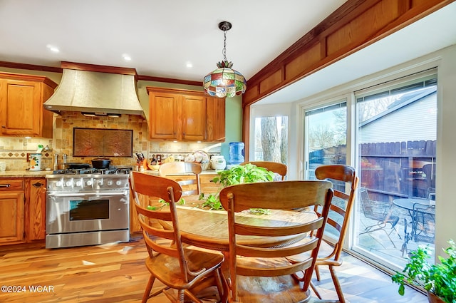 kitchen featuring stainless steel gas stove, brown cabinetry, decorative backsplash, premium range hood, and pendant lighting