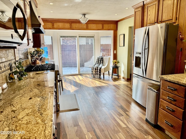 kitchen with stainless steel fridge, brown cabinetry, light stone counters, crown molding, and light wood-type flooring