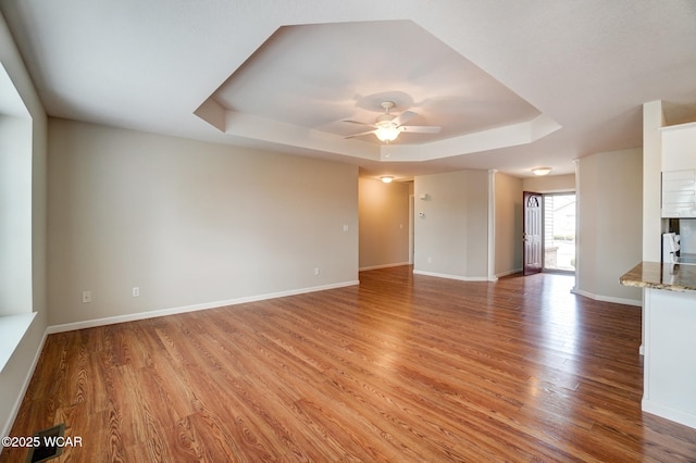 unfurnished living room featuring ceiling fan, a raised ceiling, light wood-style flooring, and baseboards