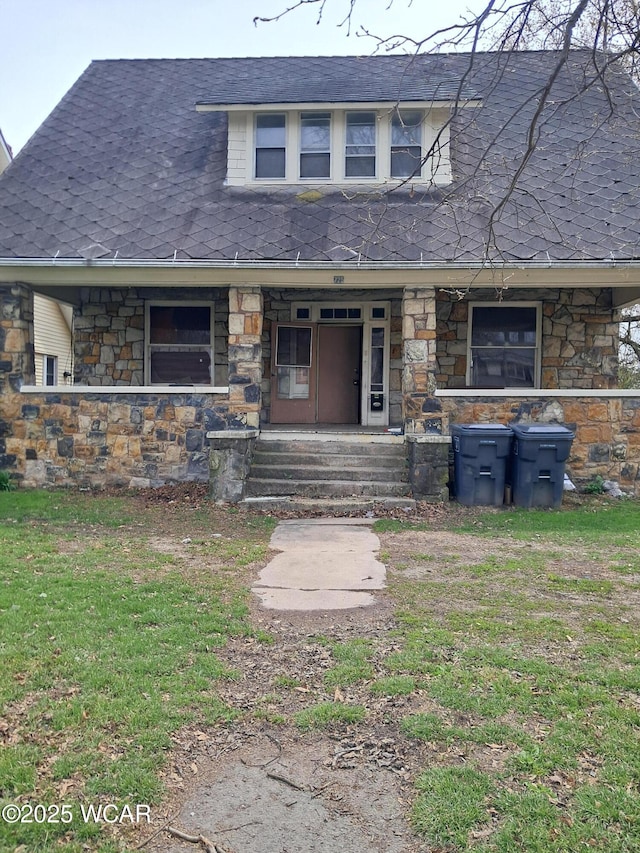 view of front facade with stone siding, a porch, roof with shingles, and a front yard