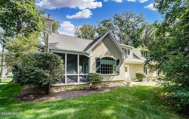 rear view of house with stucco siding, a chimney, a sunroom, a shingled roof, and a lawn