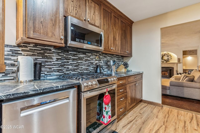 kitchen with light hardwood / wood-style flooring, a tile fireplace, appliances with stainless steel finishes, dark stone countertops, and tasteful backsplash