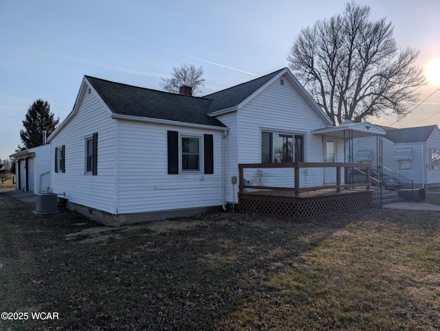 back of house at dusk with a yard, a chimney, a shingled roof, a deck, and central air condition unit