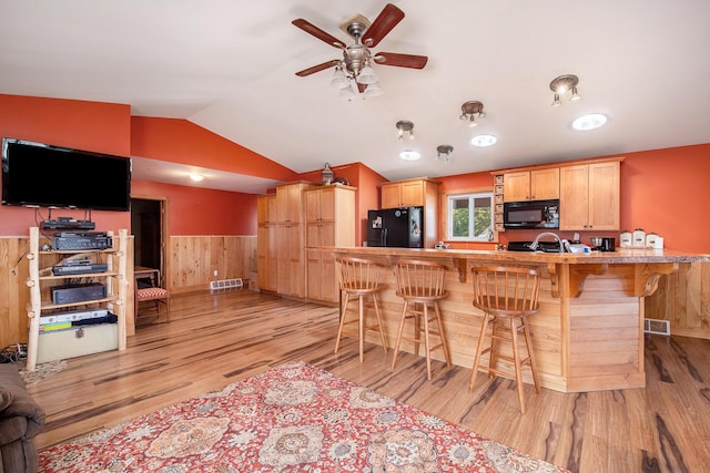 kitchen with black appliances, light brown cabinetry, a breakfast bar area, wainscoting, and lofted ceiling