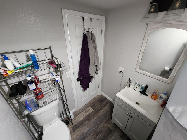 half bath with toilet, wood finished floors, baseboards, vanity, and a textured wall