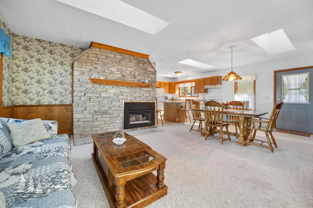 living area with wallpapered walls, a skylight, a stone fireplace, wainscoting, and light colored carpet