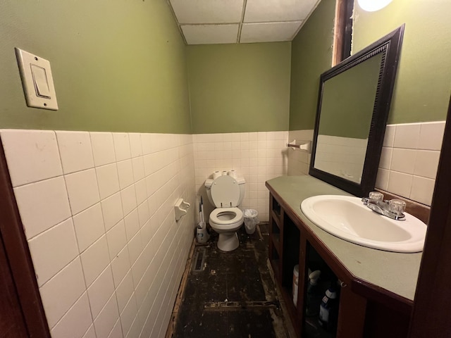 half bath with tile walls, a drop ceiling, a wainscoted wall, toilet, and vanity