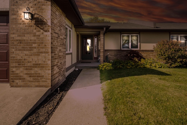 exterior entry at dusk featuring brick siding, stucco siding, and a yard