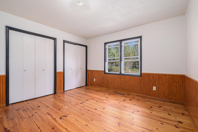 unfurnished bedroom with a wainscoted wall, a textured ceiling, two closets, and light wood-type flooring