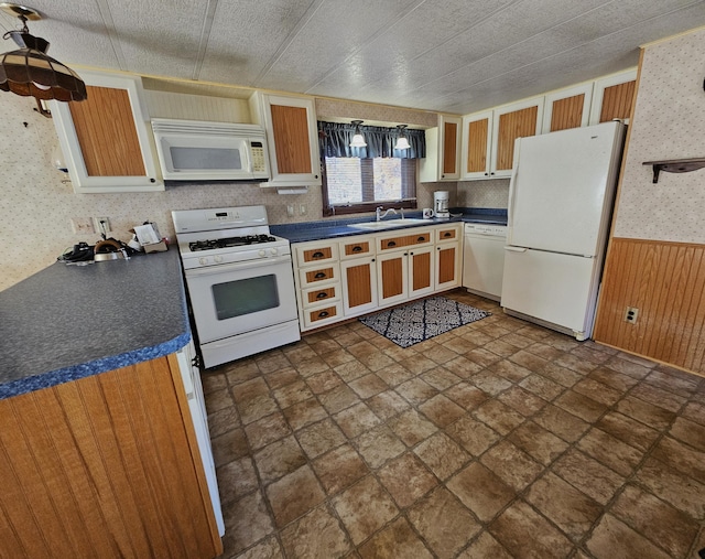 kitchen featuring wallpapered walls, a sink, wainscoting, white appliances, and dark countertops
