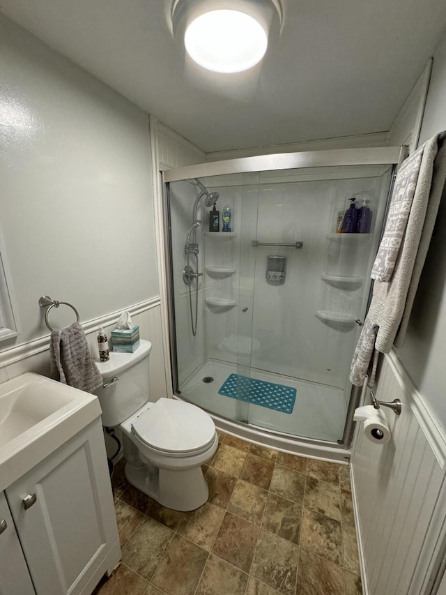 bathroom featuring vanity, a wainscoted wall, a stall shower, stone finish floor, and toilet