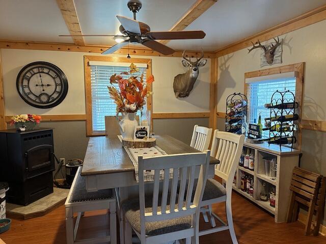 dining space featuring beam ceiling, a ceiling fan, wood finished floors, wainscoting, and a wood stove