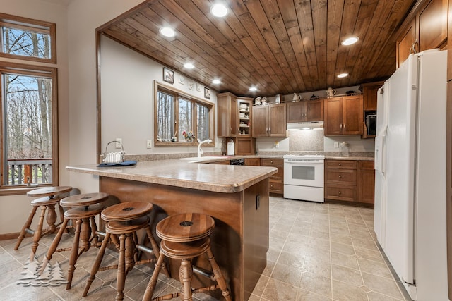 kitchen with white appliances, a peninsula, light countertops, under cabinet range hood, and wooden ceiling