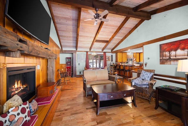 living room with a ceiling fan, lofted ceiling with beams, a lit fireplace, light wood-style floors, and wooden ceiling