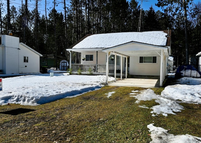 snow covered front of house featuring an outbuilding, a porch, a carport, and a shed