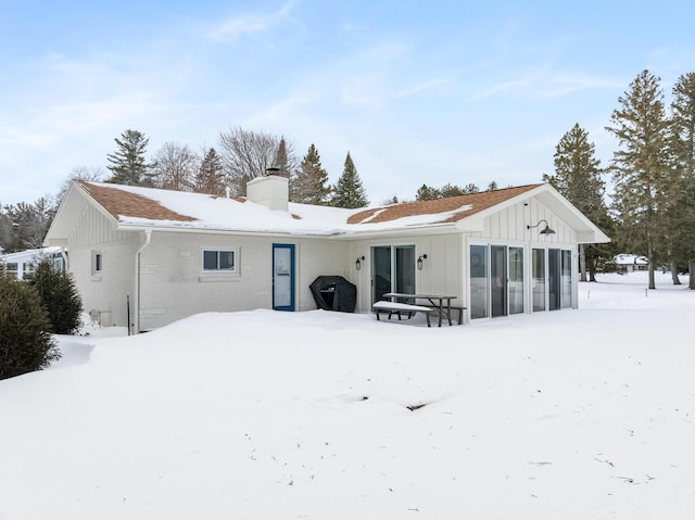 snow covered rear of property featuring brick siding, board and batten siding, and a chimney