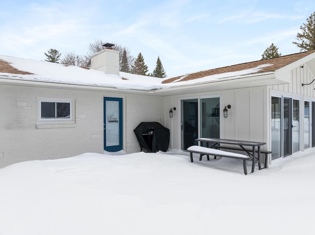 snow covered back of property with brick siding, board and batten siding, and a chimney