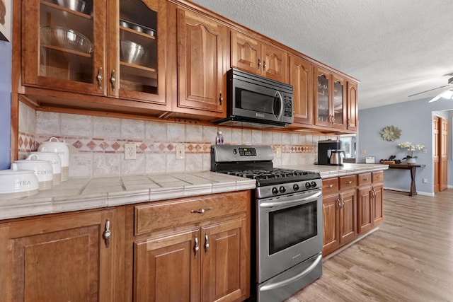 kitchen featuring brown cabinets, decorative backsplash, stainless steel appliances, light wood-style floors, and glass insert cabinets