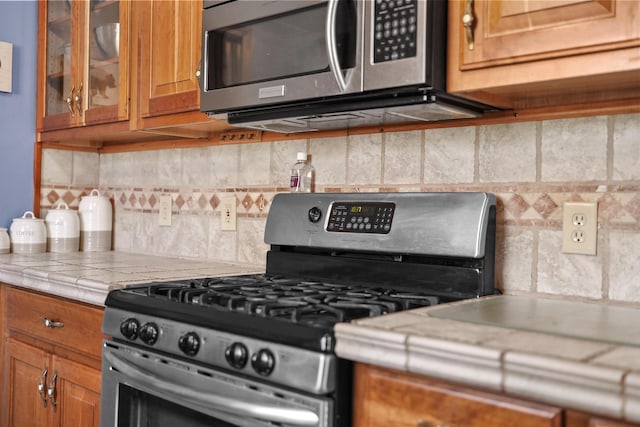 kitchen featuring tasteful backsplash, appliances with stainless steel finishes, brown cabinetry, light countertops, and glass insert cabinets