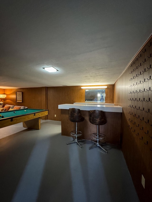 game room with a textured ceiling, pool table, concrete flooring, wood walls, and a bar