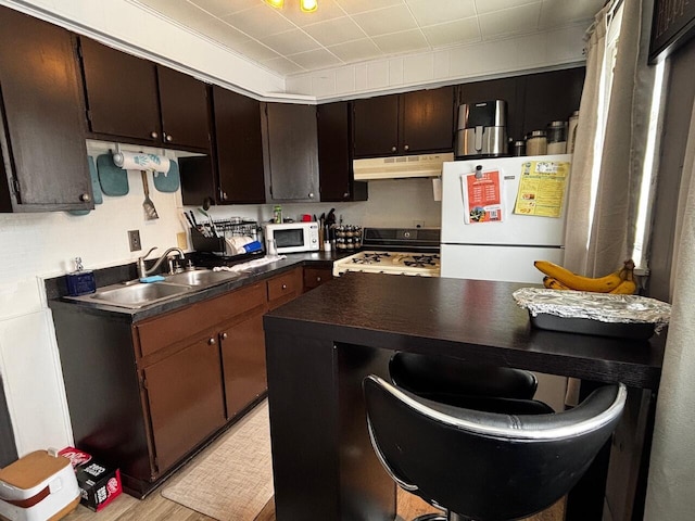 kitchen with dark countertops, dark brown cabinets, white appliances, under cabinet range hood, and a sink