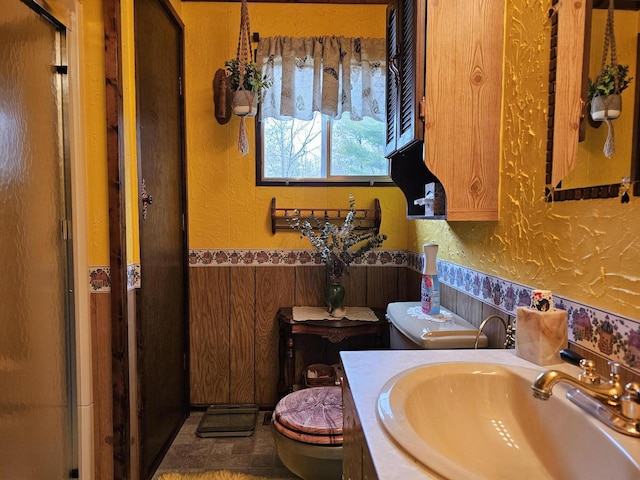 bathroom featuring toilet, vanity, a textured wall, a shower stall, and wainscoting