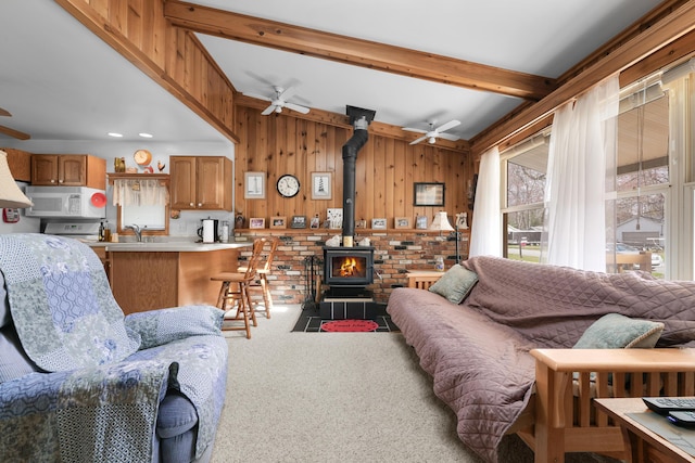 living room with wooden walls, ceiling fan, beam ceiling, a wood stove, and carpet floors