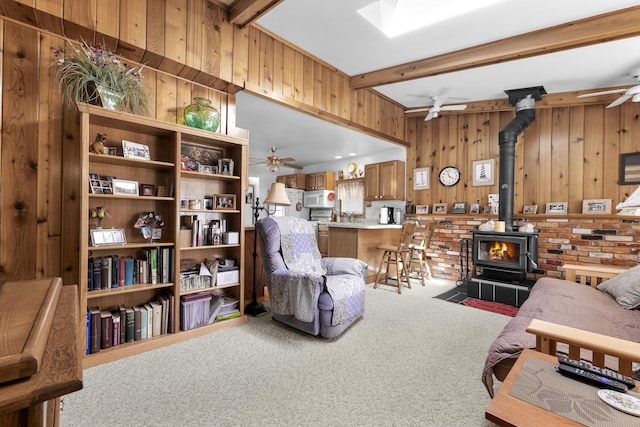 carpeted living area featuring wooden walls, ceiling fan, beamed ceiling, and a wood stove
