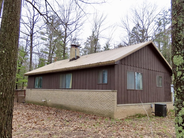 view of property exterior featuring a chimney, brick siding, central AC unit, board and batten siding, and metal roof