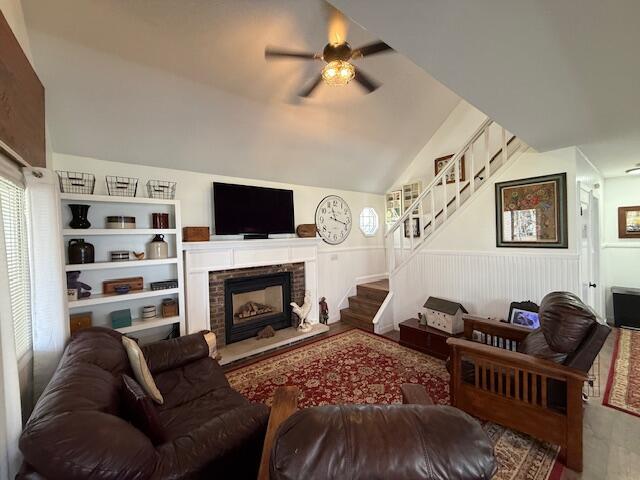 living room featuring stairway, ceiling fan, wainscoting, a fireplace with raised hearth, and vaulted ceiling
