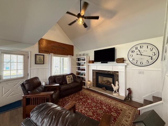 living area with wainscoting, a ceiling fan, wood finished floors, a fireplace with raised hearth, and vaulted ceiling