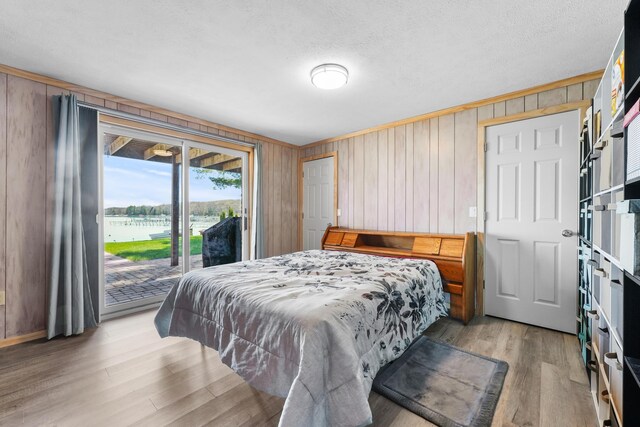 bedroom with access to exterior, light wood-type flooring, wood walls, a water view, and a textured ceiling