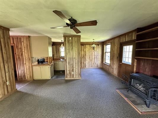 605 N Poor Farm Rd, Harrisville, MI 48740 - unfurnished living room with a wood stove, wooden walls, healthy amount of natural light, ceiling fan, and carpet floors