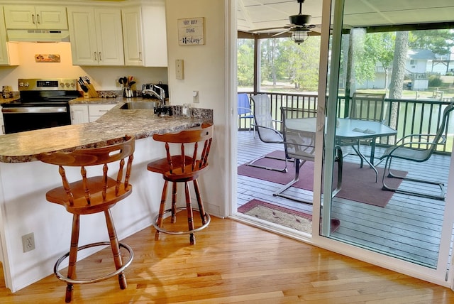 kitchen with stainless steel electric range oven, ventilation hood, a breakfast bar area, ceiling fan, and light wood-type flooring