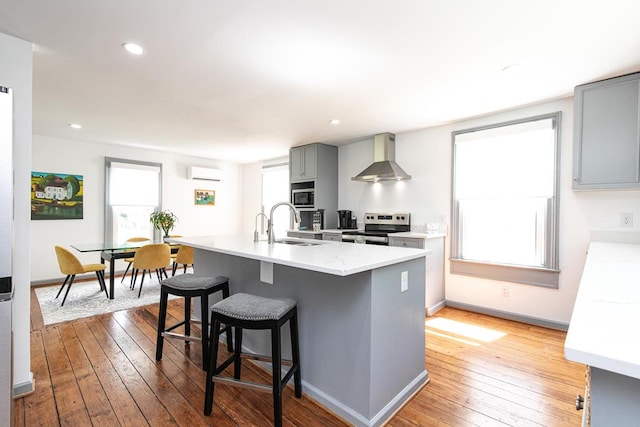 kitchen featuring wall chimney exhaust hood, gray cabinetry, a center island with sink, an AC wall unit, and stainless steel electric stove