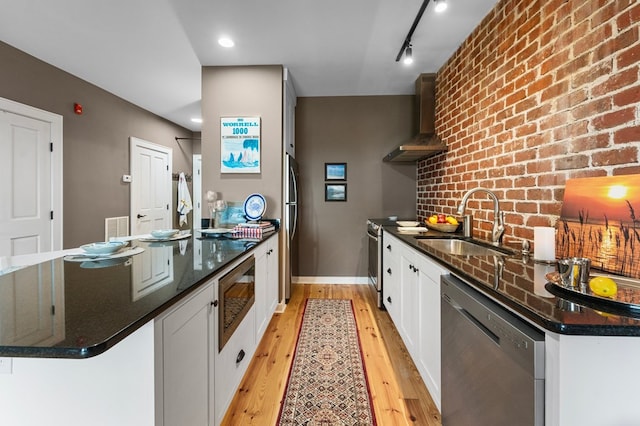 kitchen with light wood-style flooring, appliances with stainless steel finishes, wall chimney range hood, white cabinetry, and a sink