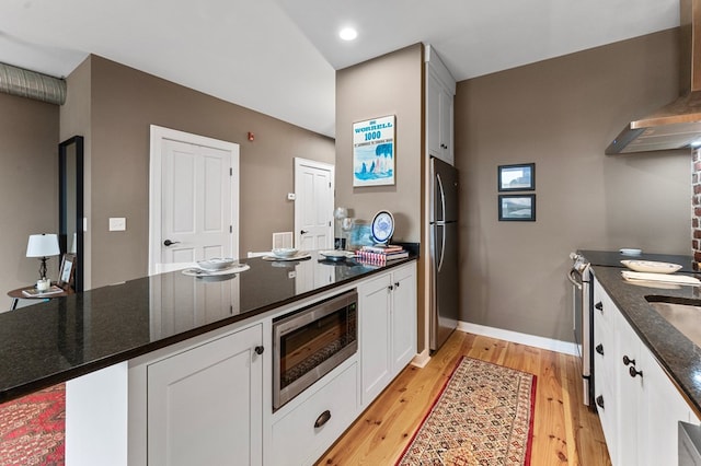 kitchen with wall chimney range hood, appliances with stainless steel finishes, white cabinetry, dark stone countertops, and light wood-type flooring