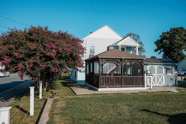 view of front facade featuring an outbuilding, roof with shingles, a gazebo, a shed, and a front lawn