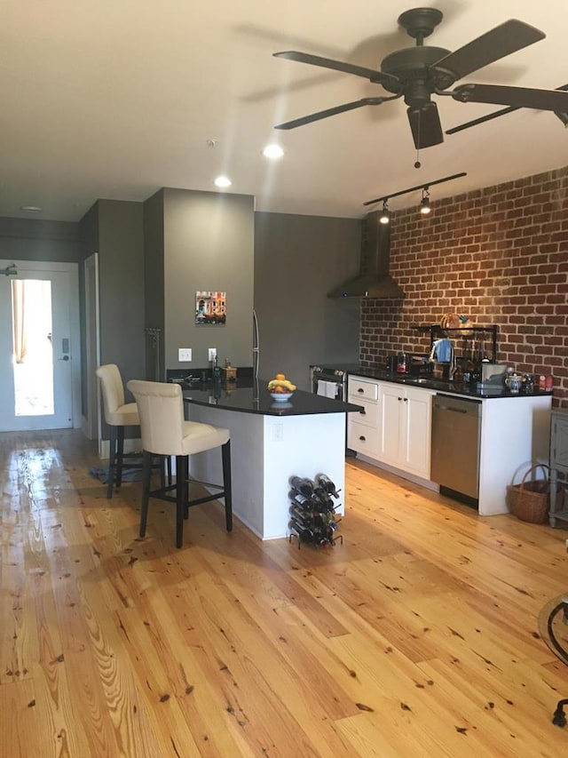 kitchen featuring wall chimney range hood, white cabinetry, stainless steel dishwasher, dark countertops, and light wood-type flooring
