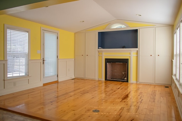 unfurnished living room featuring lofted ceiling, a decorative wall, a wainscoted wall, a fireplace, and light wood finished floors