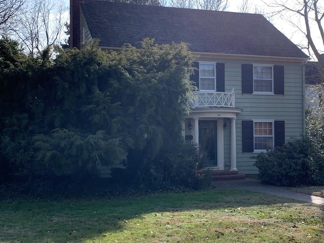 view of front facade featuring a balcony, a shingled roof, a chimney, and a front yard