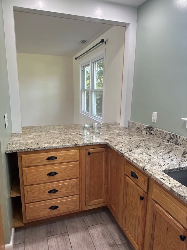 kitchen with brown cabinetry, visible vents, a peninsula, light wood-style floors, and light stone counters