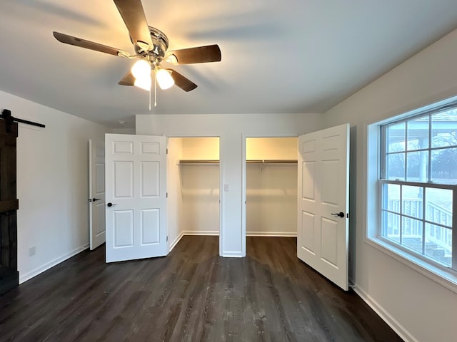 unfurnished bedroom featuring two closets, dark wood-type flooring, a barn door, and ceiling fan