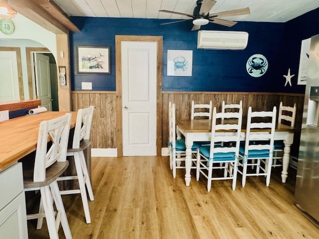 dining room featuring a wall mounted air conditioner, a ceiling fan, wainscoting, wooden walls, and light wood-style flooring