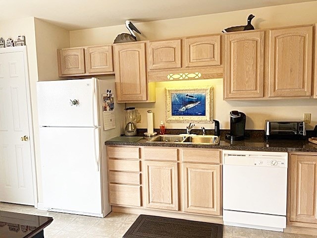 kitchen with light brown cabinetry, white appliances, dark countertops, and a sink