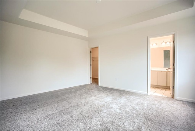 unfurnished bedroom with a walk in closet, a raised ceiling, light carpet, ensuite bath, and baseboards