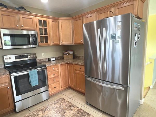 5061 Bay Colony Ln, Chincoteague, VA 23336 - kitchen featuring appliances with stainless steel finishes, light tile patterned flooring, glass insert cabinets, brown cabinetry, and dark stone countertops